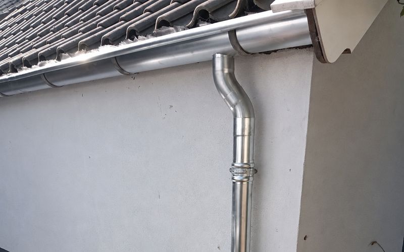 Zinken dakgoot met regenpijp en bocht langs een witte gevel